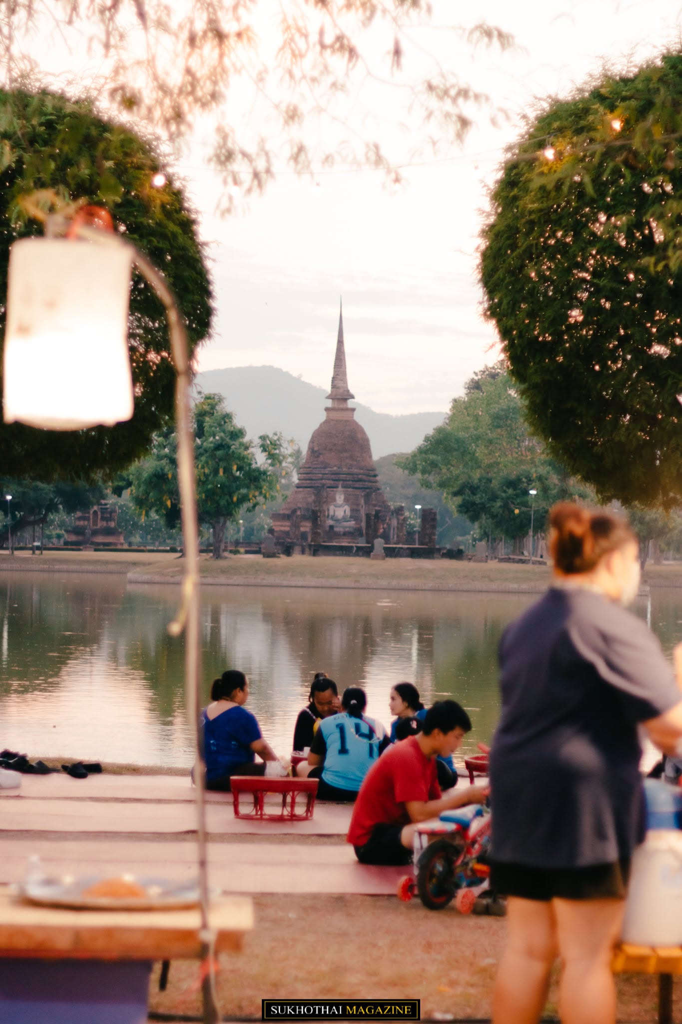 Diners seated at the lakeside khantoke area looking across the moat toward the illuminated spire of Wat Sa Si at dusk