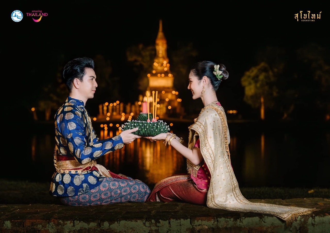 Couple in traditional Thai court dress holding a krathong by a candlelit moat at Sukhothai Historical Park
