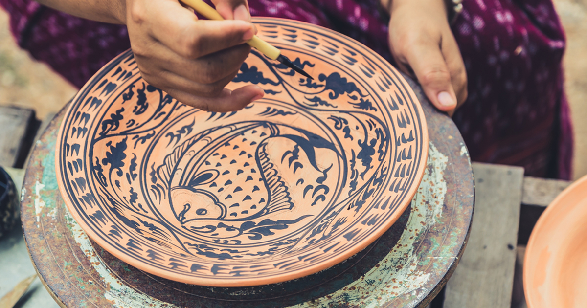 Artisan painting an elaborate kadam fish and botanical scroll pattern on a terracotta plate