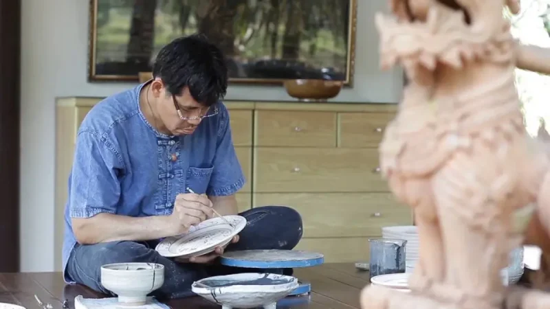 Artisan painting a celadon plate in a serene studio workshop, surrounded by finished ceramic pieces